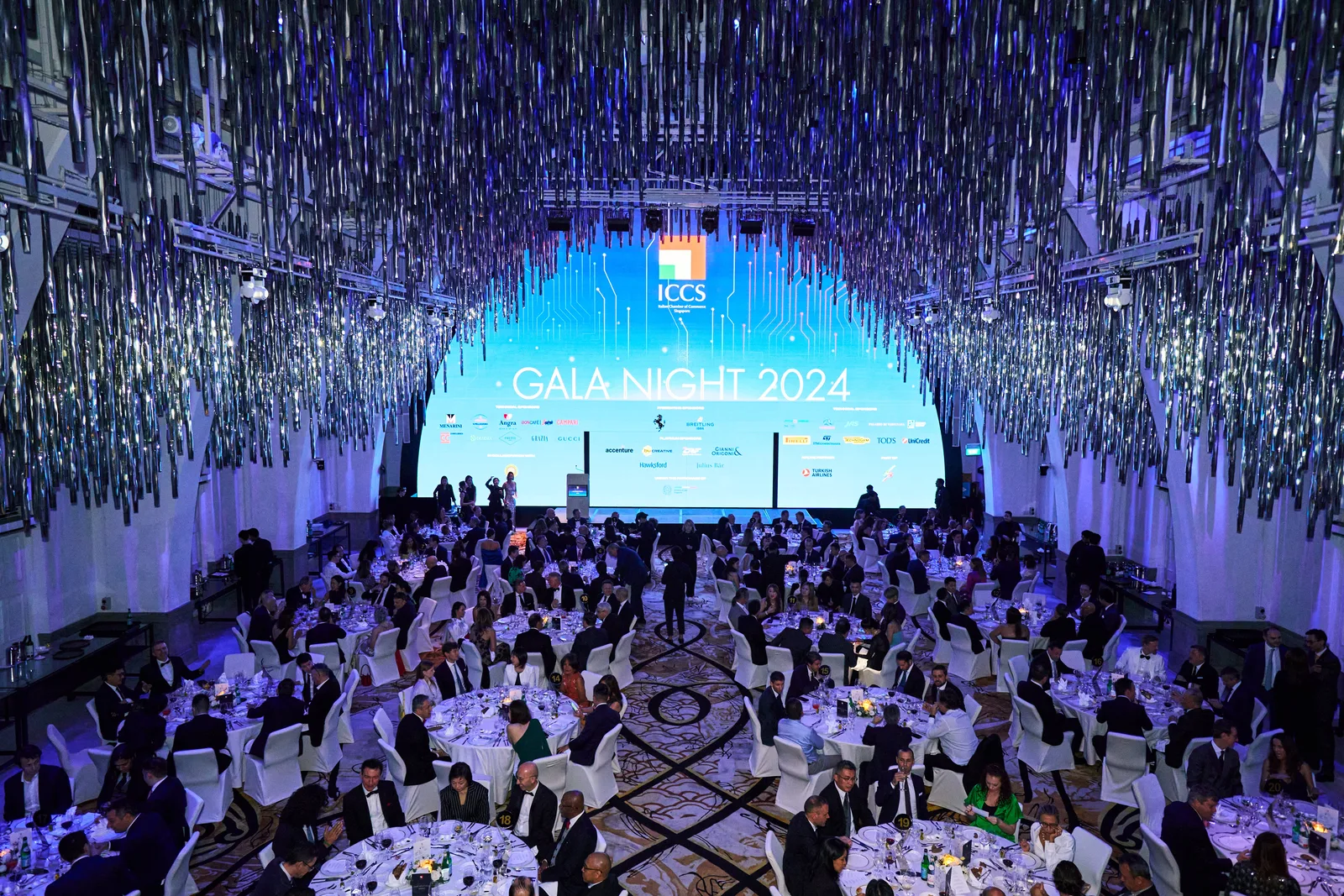 Wide gala ballroom under hanging silver streamers with a lit stage screen above full banquet tables