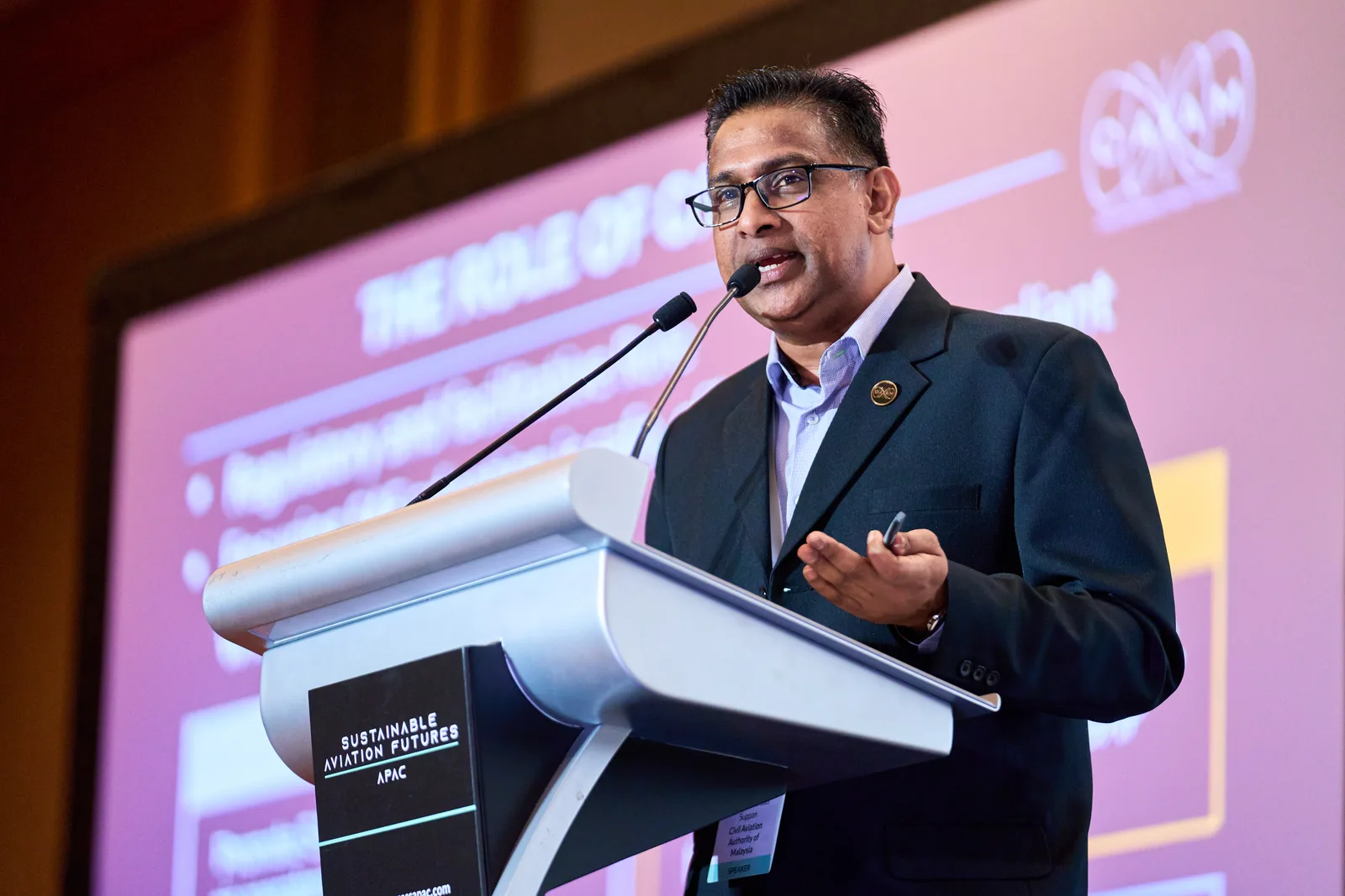 Speaker delivering a keynote from a podium beside a presentation screen at a Singapore conference