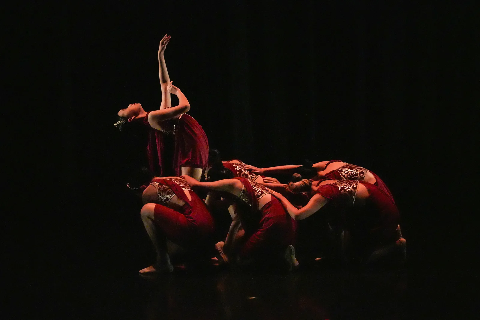 Contemporary dancers grouped on a dark stage under deep red lighting