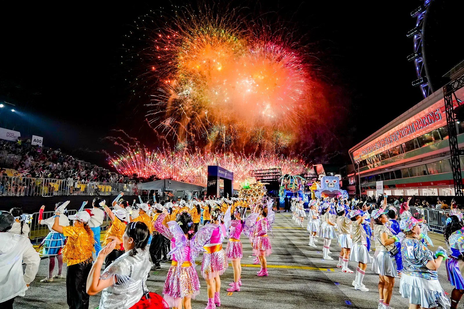 Fireworks bursting over a night parade ground with costumed performers and a packed grandstand, Singapore