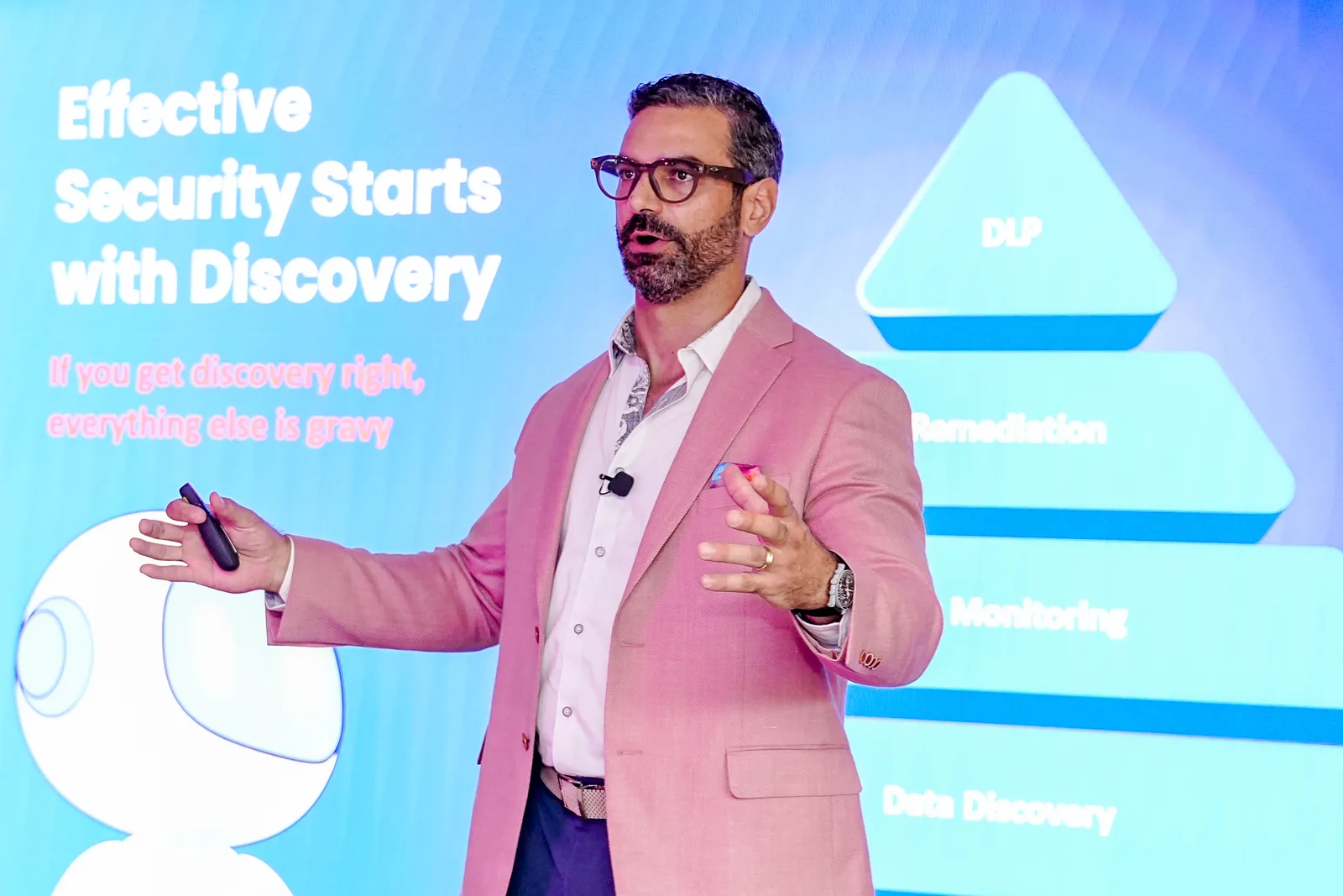 Conference keynote speaker mid-gesture on stage in front of a large blue presentation screen